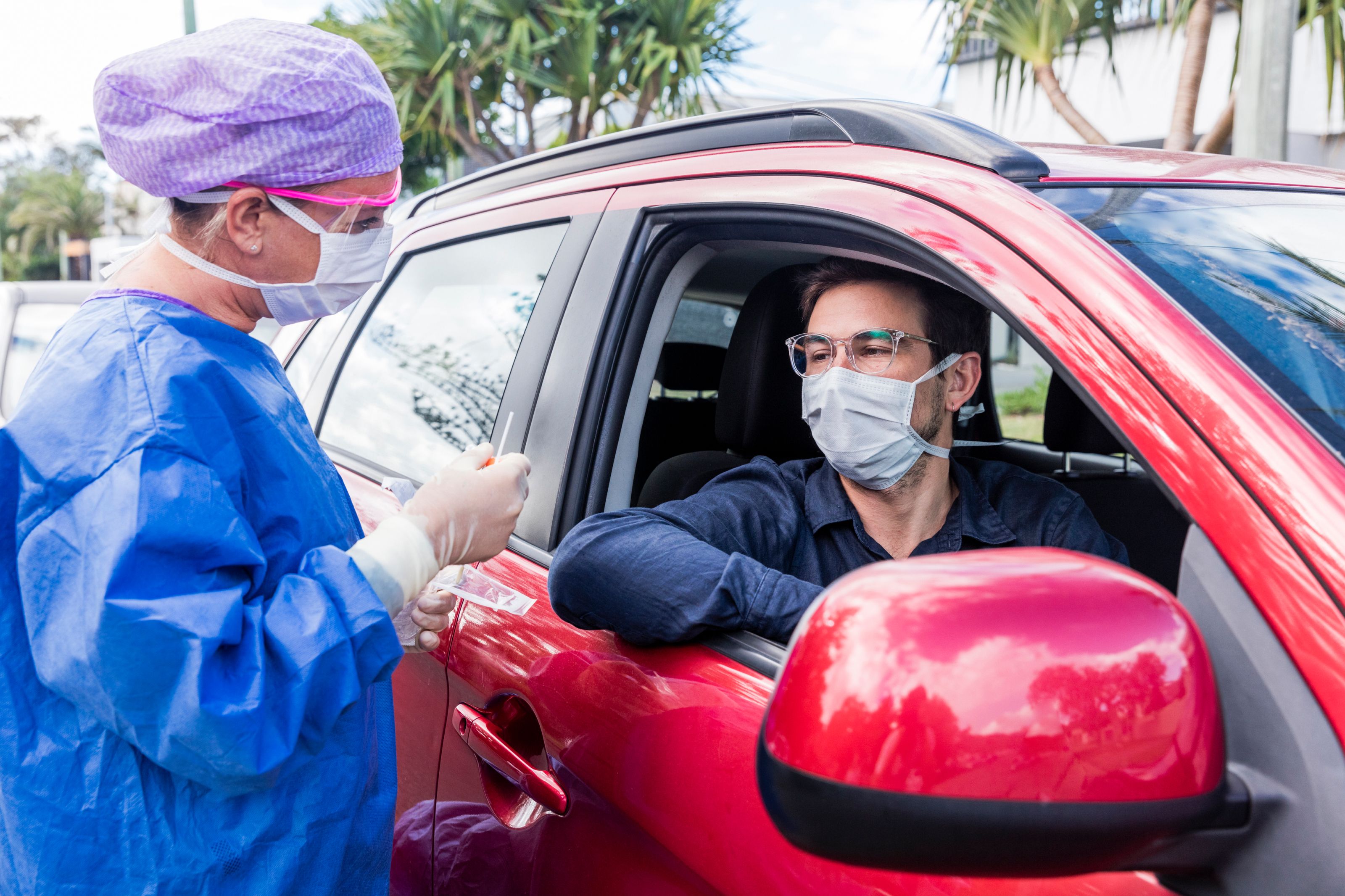 man in red car wearing mask receiving rapid covid test from nurse in blue protective gown